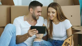 Couple sitting on the floor with moving boxes, using a tablet while planning their home move