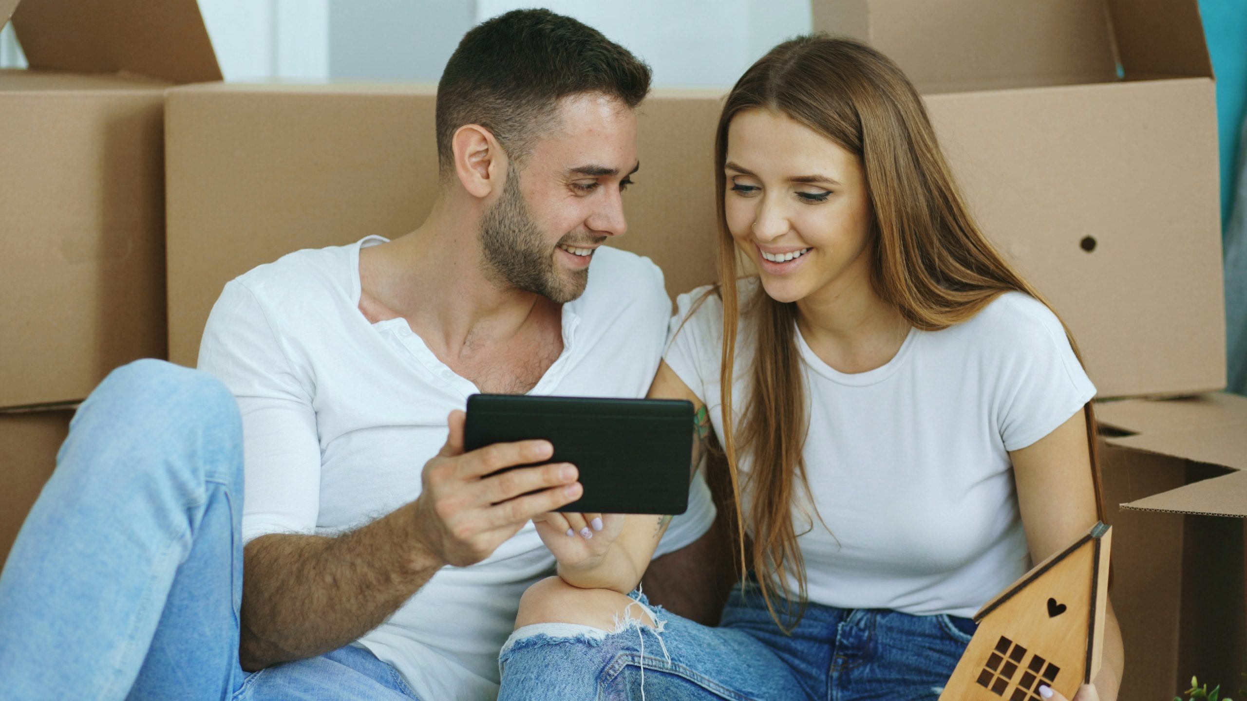 Couple sitting on the floor with moving boxes, using a tablet while planning their home move